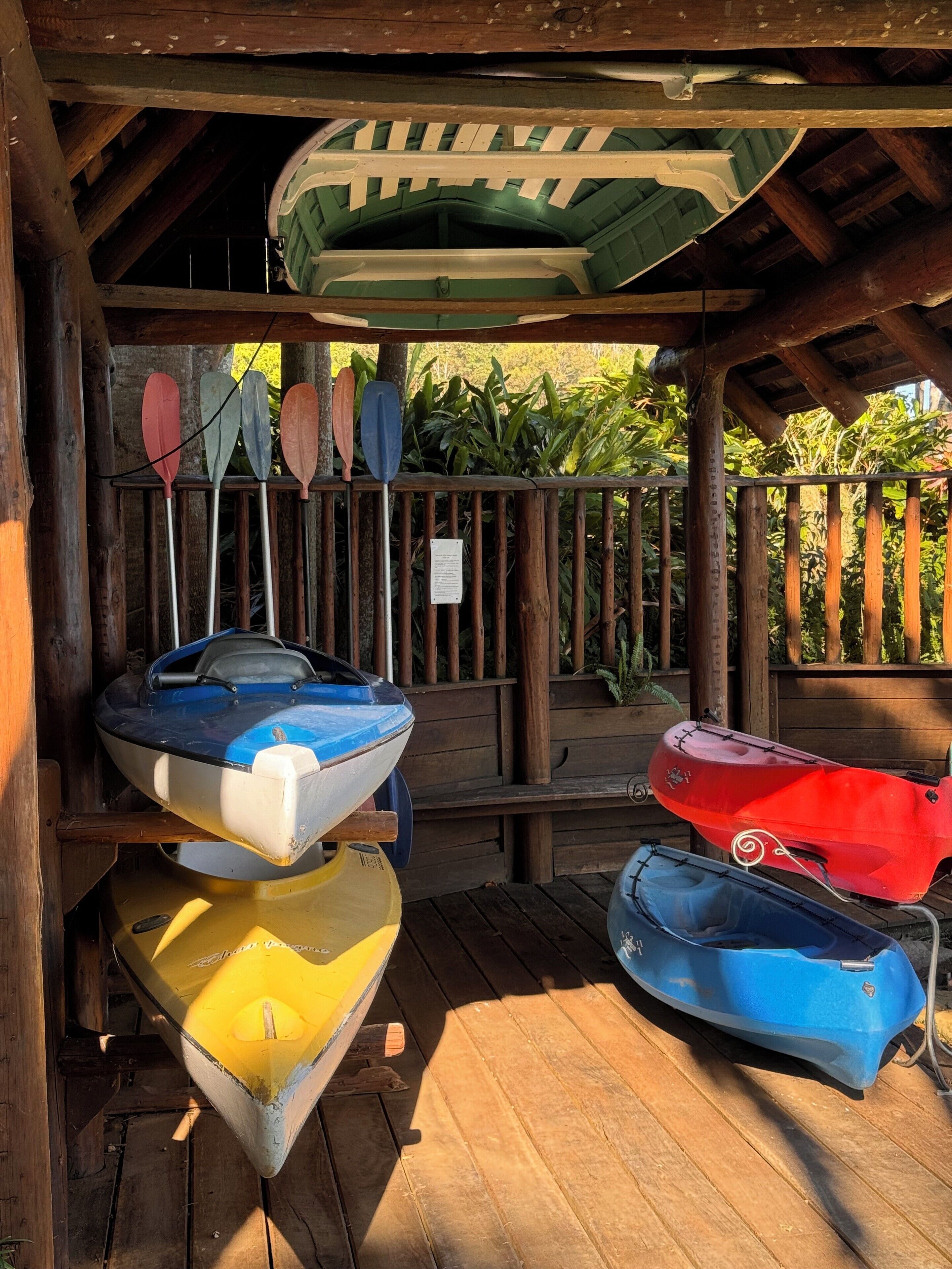 Une terrasse en bois couverte, sous laquelle sont rangés plusieurs kayaks et planches de paddle.