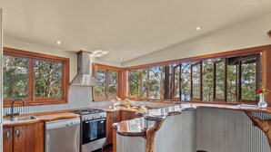 A modern kitchen with wooden cabinets, stainless steel appliances, and large windows offering a view of trees. - Secrets on the Lake (North Maleny)