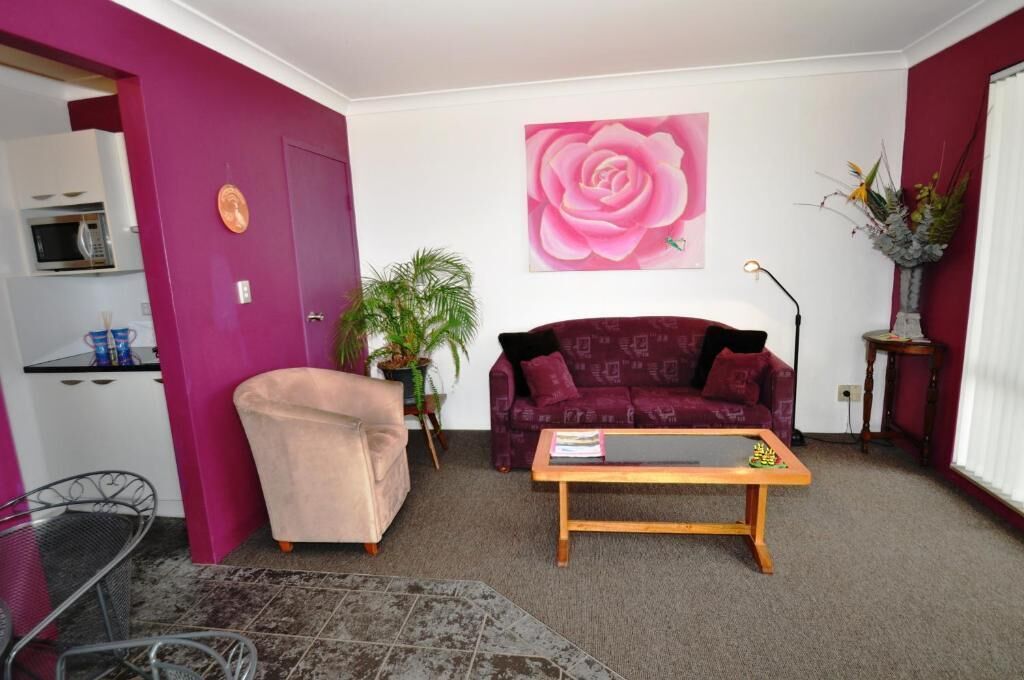 A living room with a purple sofa, a pink flower painting, a coffee table, and a small kitchen area with a microwave.