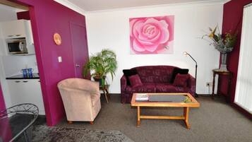 A living room with a purple sofa, a pink flower painting, a coffee table, and a small kitchen area with a microwave.