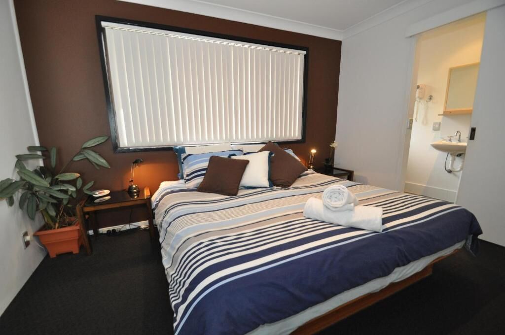 Een hotelkamer met een bed, nachtkastje, lamp, kamerplant en een raam met jaloezieën.