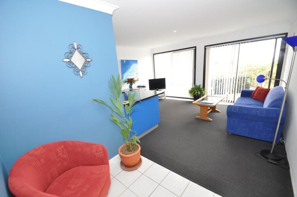 A modern living room with a blue sofa, a red chair, a TV, and a balcony with large windows.