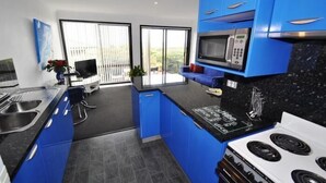A modern kitchen with blue cabinets, a microwave, a stove, and a dining area with a TV and sofa. - Beachfront Apartments Narooma (North Narooma)