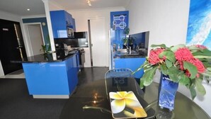 A modern hotel lobby with a blue reception desk, a glass dining table with a floral placemat, and a vase with red flowers. - Beachfront Apartments Narooma (North Narooma)