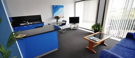 A modern hotel room with a blue accent wall, a kitchenette, a seating area with a sofa and a coffee table, and large windows with blinds.