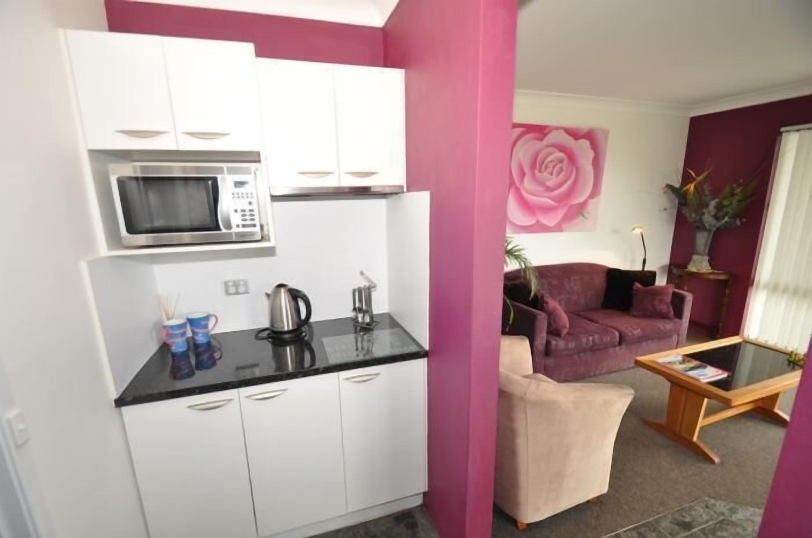 A compact living space with a kitchenette, a pink sofa, a coffee table, and a large floral painting.