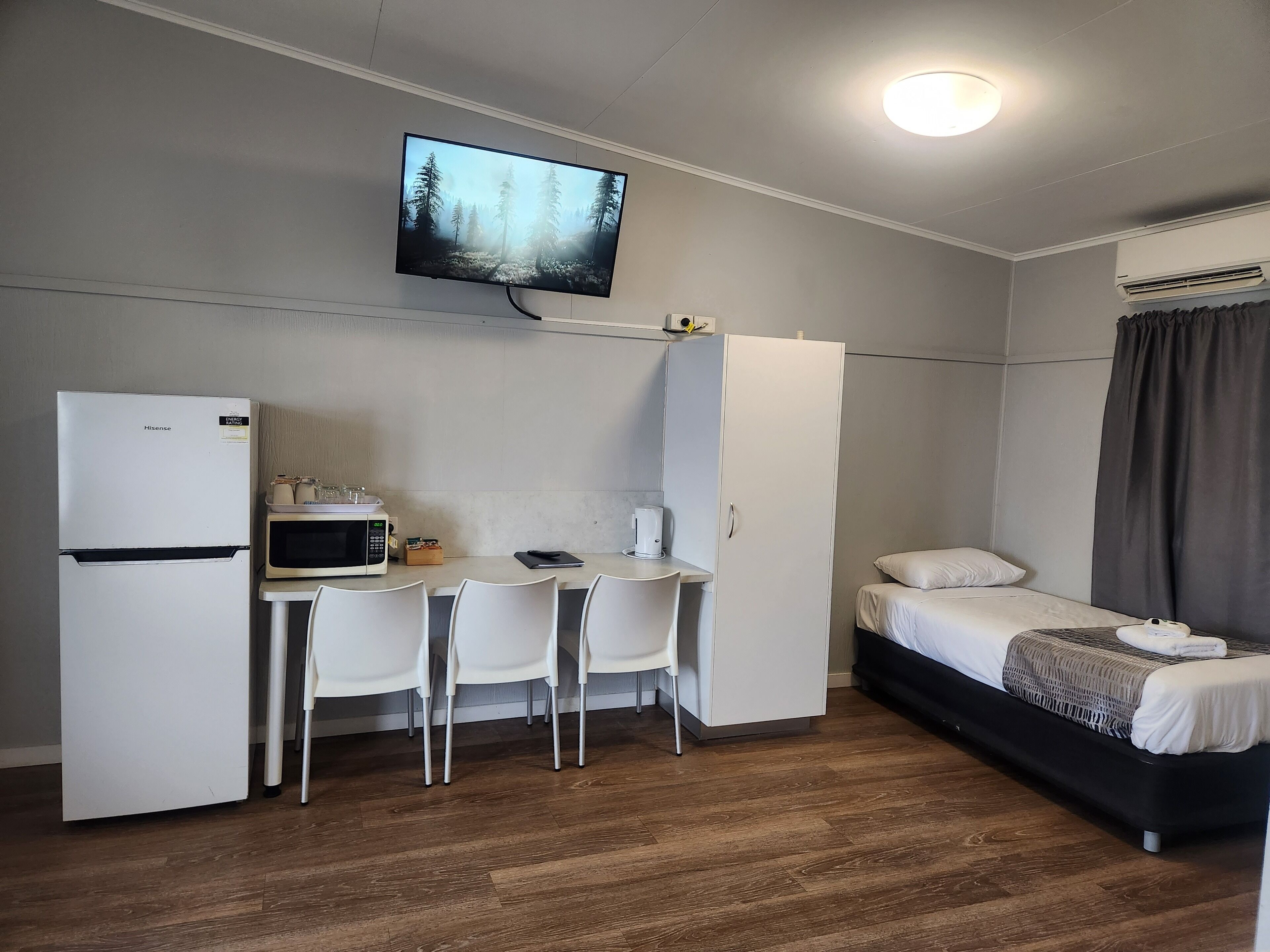 Quarto de hotel compacto com uma cama, escrivaninha com cadeira, micro-ondas, frigorífico, televisão e guarda-roupa.