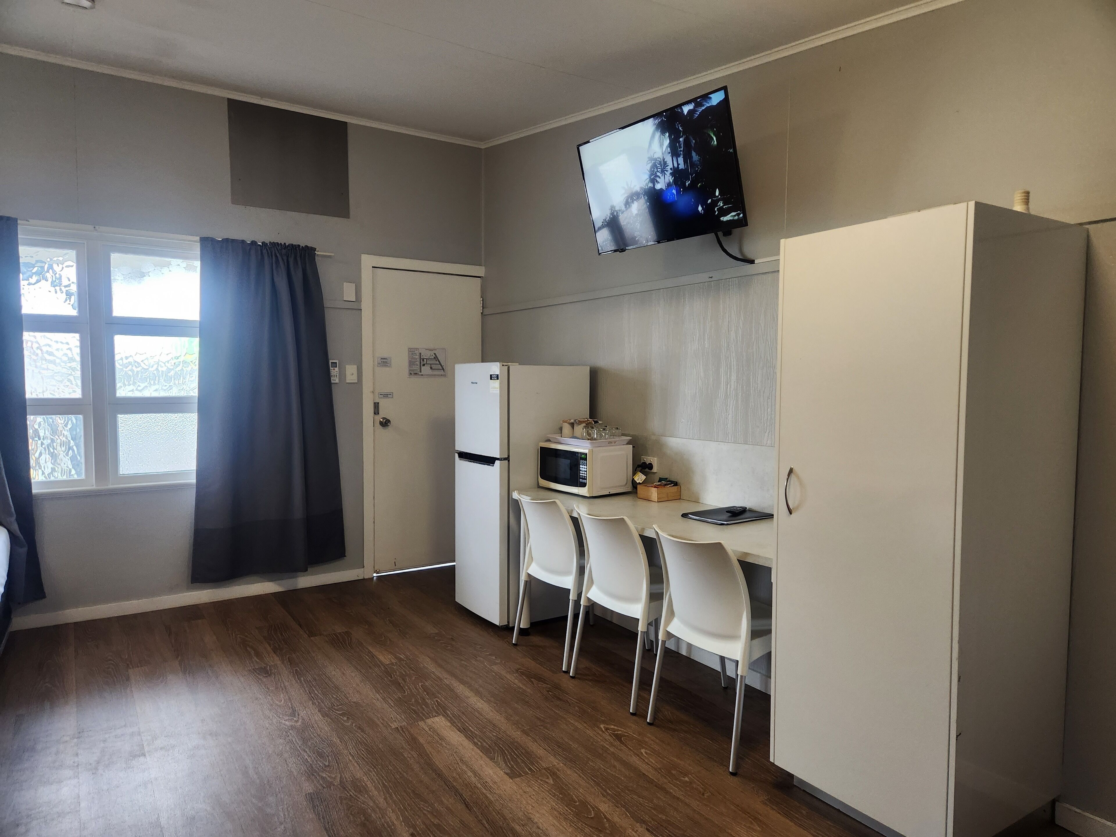 A compact kitchen with a refrigerator, microwave, and a TV mounted on the wall. There are three white chairs and a small table.