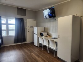 Cocina compacta con refrigerador, microondas y televisión instalada en la pared. Hay tres sillas blancas y una mesa pequeña.