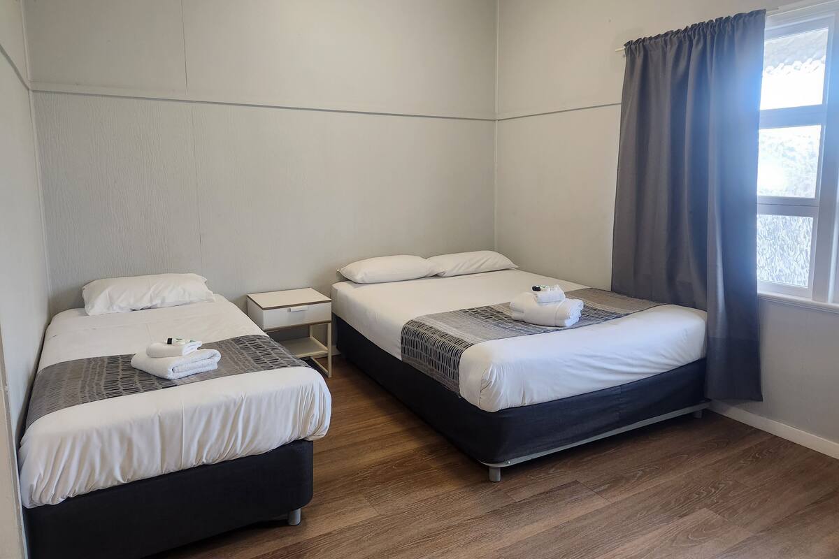 Hotellihuone, jossa on kaksi sänkyä, pieni yöpöytä, ikkuna verhoilla ja puulattia.
