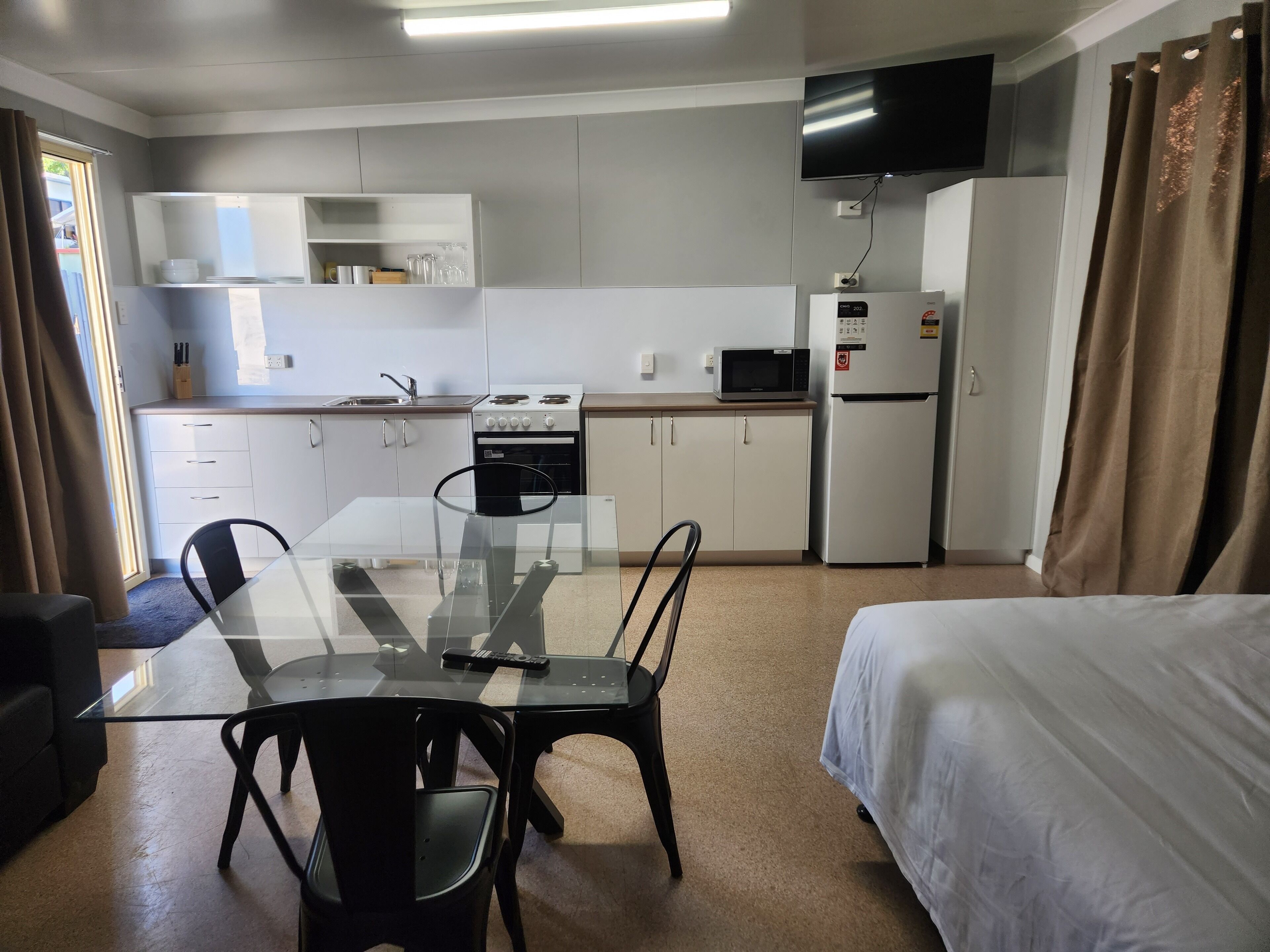 A compact hotel room with a kitchenette, a dining table with chairs, a bed, and a TV mounted on the wall.