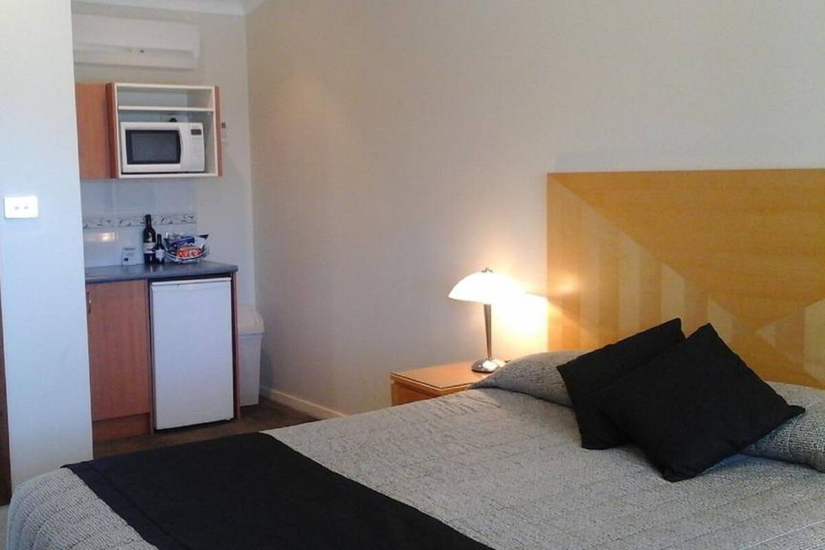 Habitación de hotel con cama, una lámpara de noche, una kitchenette pequeña con microondas y vista a la habitación contigua.