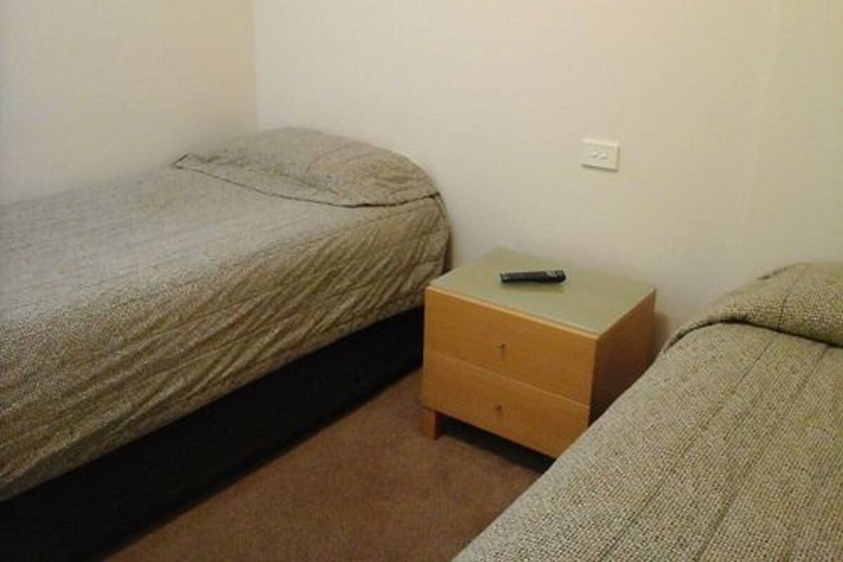 Una habitación pequeña y sencilla con dos camas individuales, una mesita de noche de madera y un enchufe en la pared.