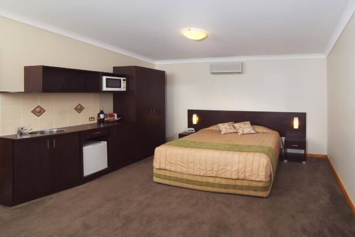 Una habitación de hotel compacta con cama, mesitas de noche, una kitchenette pequeña y un microondas.