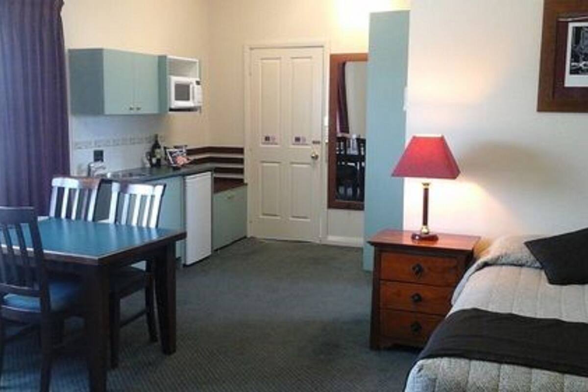 Habitación de hotel pequeña con mesa de comedor, sillas, una kitchenette, cama, mesita de noche y lámpara.
