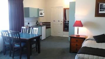 A compact hotel room with a dining table, chairs, a small kitchenette, a bed, a nightstand, and a lamp.
