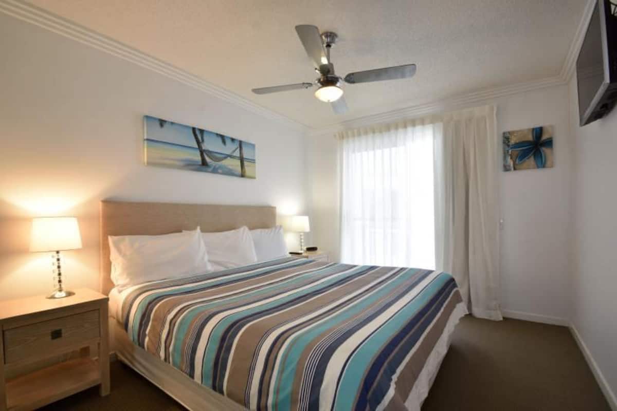 Une chambre d’hôtel avec un lit, deux lampes de chevet, un ventilateur de plafond, un tableau et un téléviseur fixé au mur.