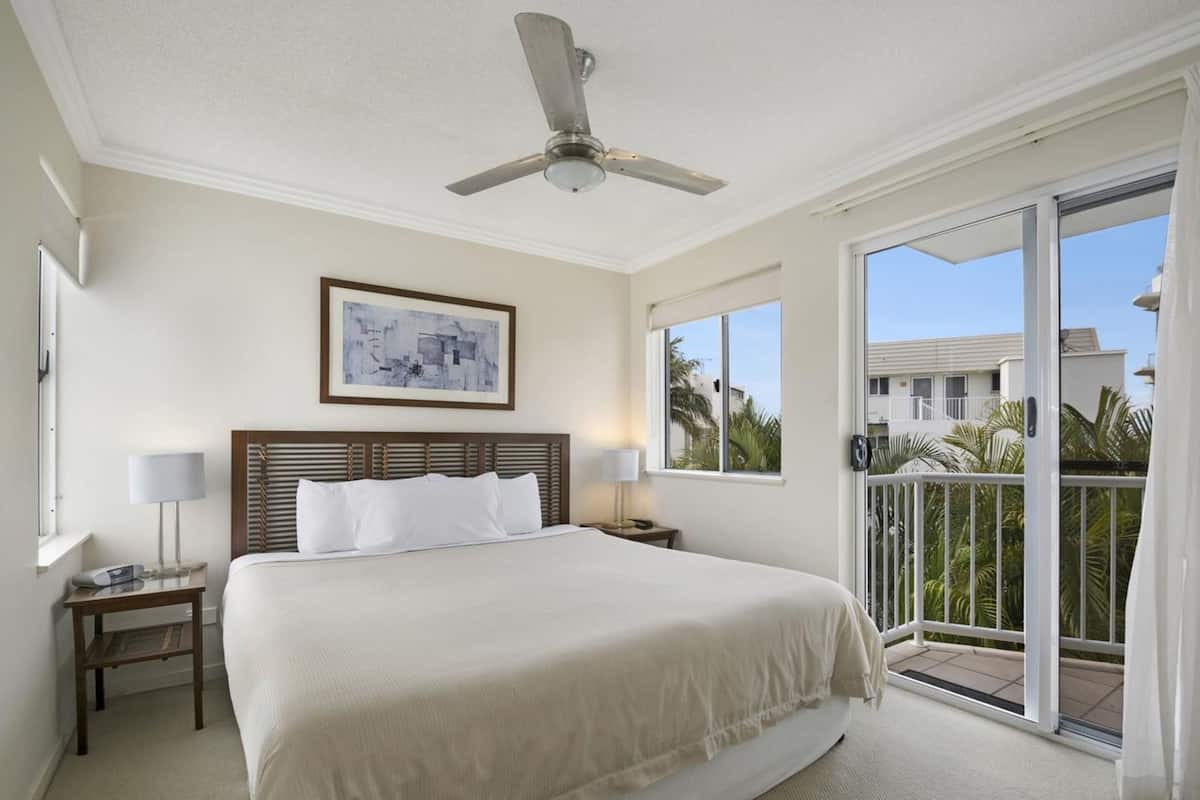 Une chambre d’hôtel avec un grand lit, un ventilateur de plafond, un tableau au mur et un balcon donnant sur des palmiers.