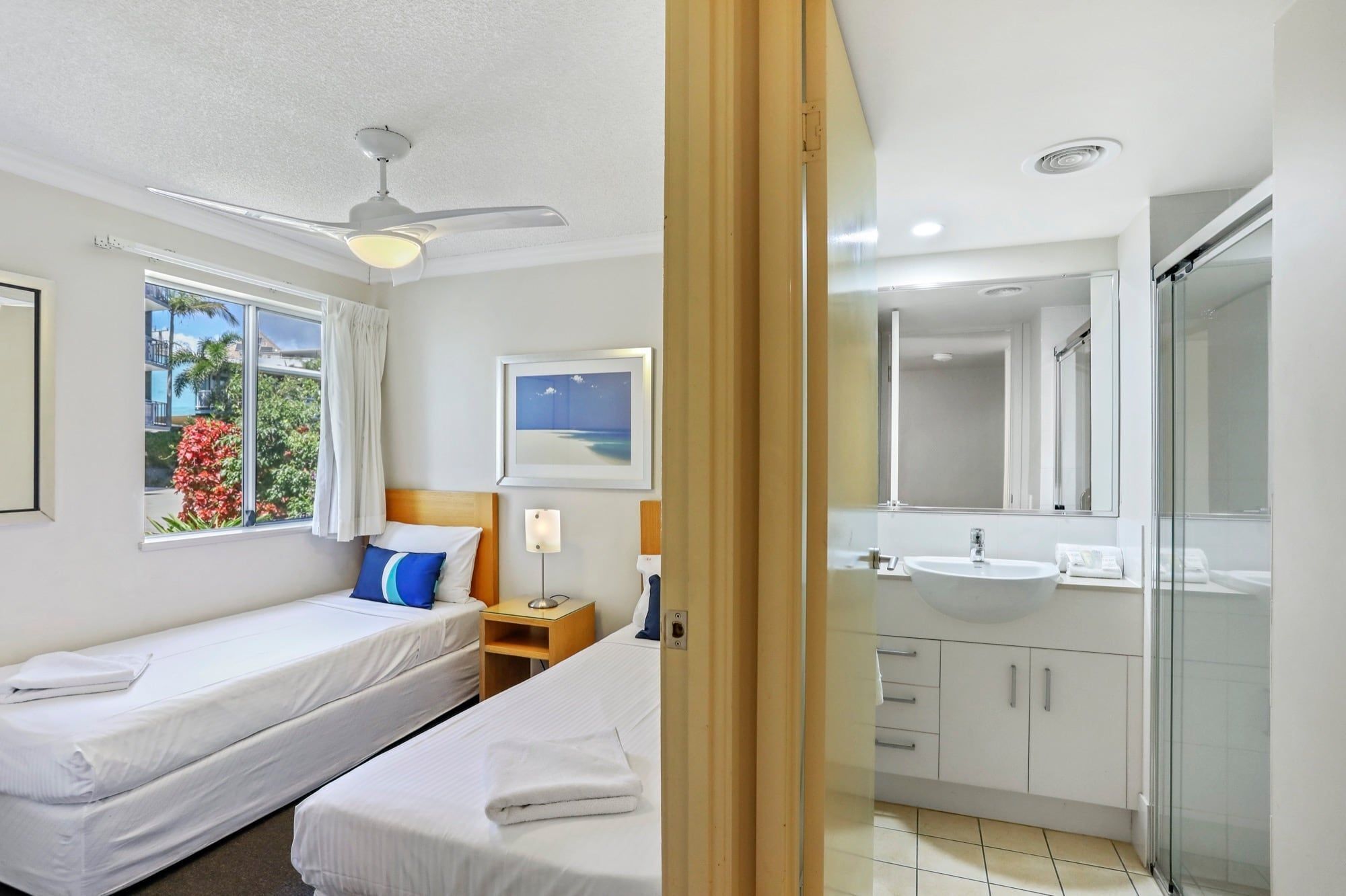 A hotel room with two beds, a ceiling fan, a window with a view of greenery, a bathroom with a sink and mirror, and a shower.