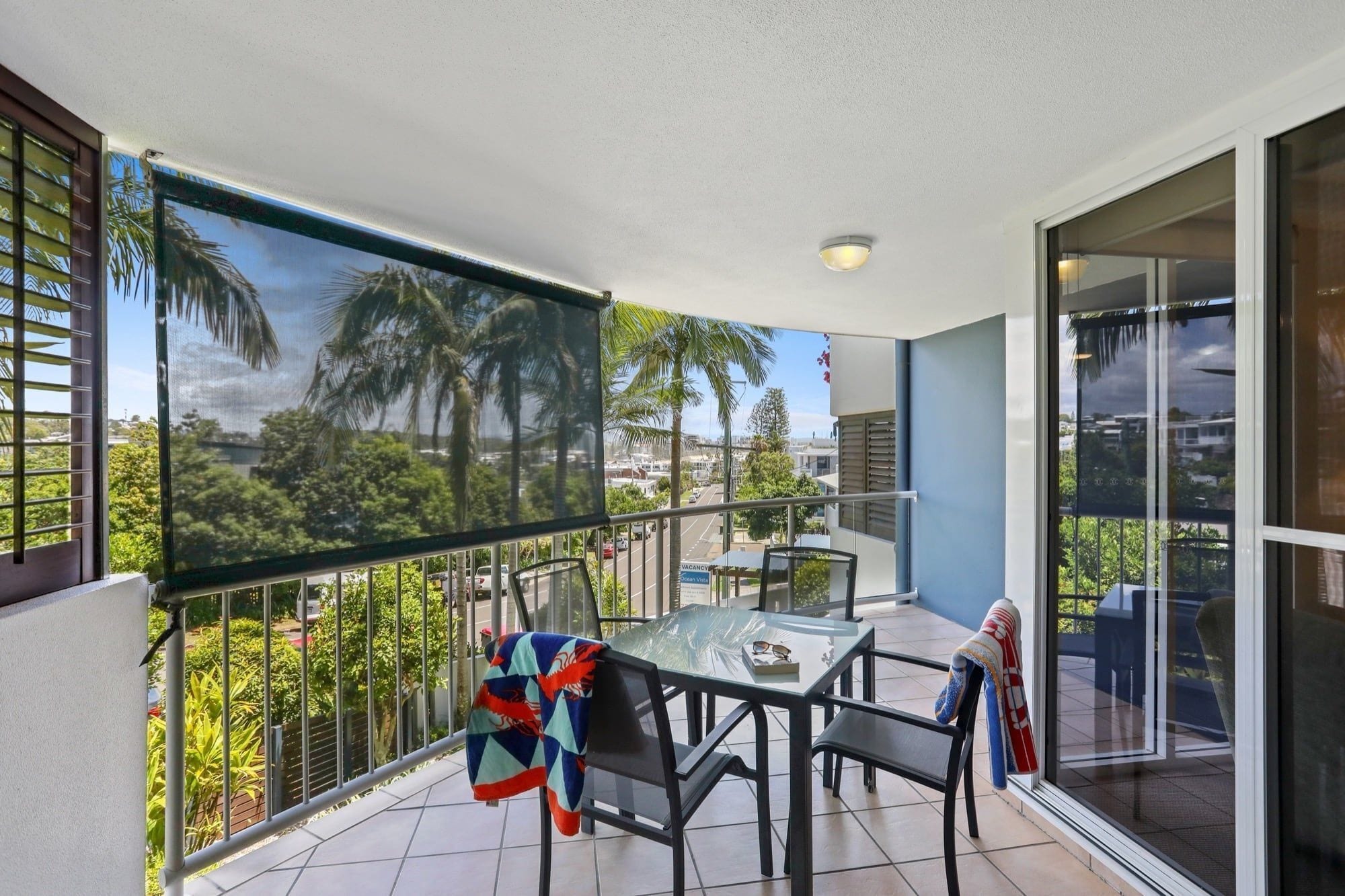 A balcony with a glass door, a table, chairs, and a view of palm trees and buildings.