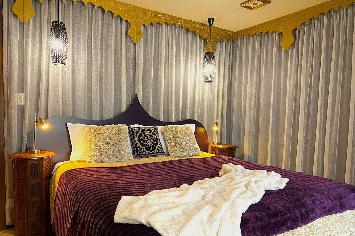 Un lit avec une couvre-lit aux motifs violets et blancs, deux lampes de chevet et une tête de lit décorative.