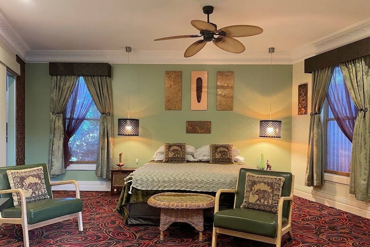 Une chambre à coucher comprenant un lit, deux chaises, une table ronde, un ventilateur de plafond et un tapis à motifs.