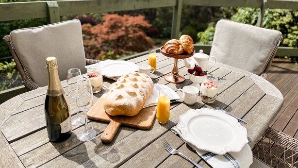 A wooden deck with a table set for breakfast, including croissants, strawberries, and a bottle of champagne. - Woolrich Historic Garden Cottage (Olinda)