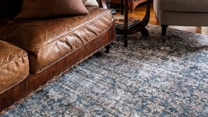 A living room with a brown leather sofa, a white armchair, a wooden cabinet, and a patterned rug. - Woolrich Historic Garden Cottage (Olinda)