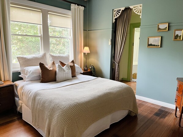 Cama bem arrumada com manta branca e bege, mesa de cabeceira com abajur e vista para o verde pela janela.