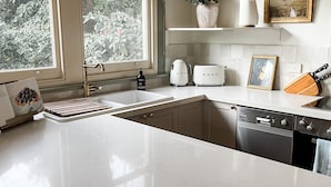 A modern kitchen with white countertops, a built-in dishwasher, a sink, and a window with a view of greenery. - Woolrich Historic Garden Cottage (Olinda)