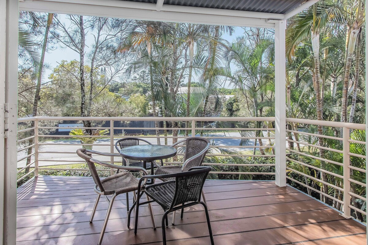 A covered outdoor seating area with metal chairs and a table, surrounded by a white railing, overlooking a road and lush greenery.