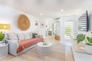A modern living room with a sofa, round coffee table, and decorative items. There is a TV mounted on the wall and a window with blinds. - Hastings Cove Waterfront Holiday Apartments (Hastings Point)