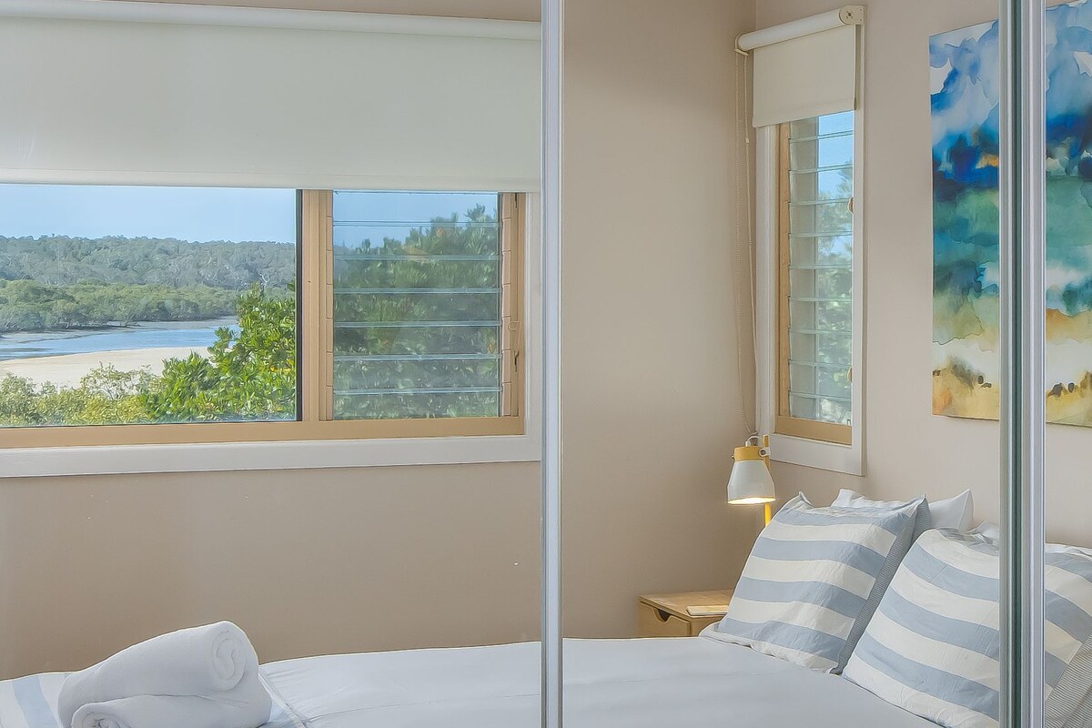 A bedroom with a bed, a nightstand, a lamp, and a view of a beach through a window.