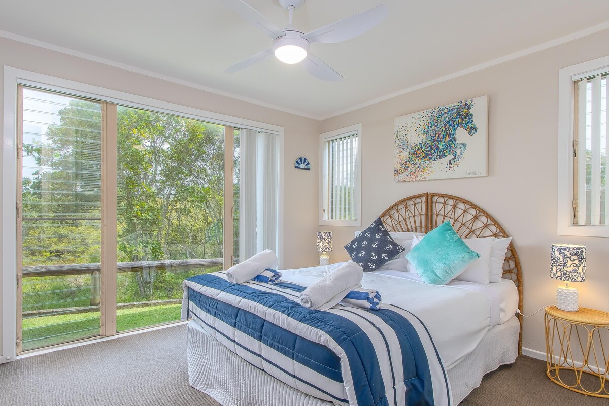A bedroom with a large bed, a ceiling fan, and a view of greenery through the windows.