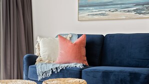 Un canapé bleu foncé avec trois coussins, un pouf en rotin tressé et un tableau représentant une scène de plage accroché au mur.