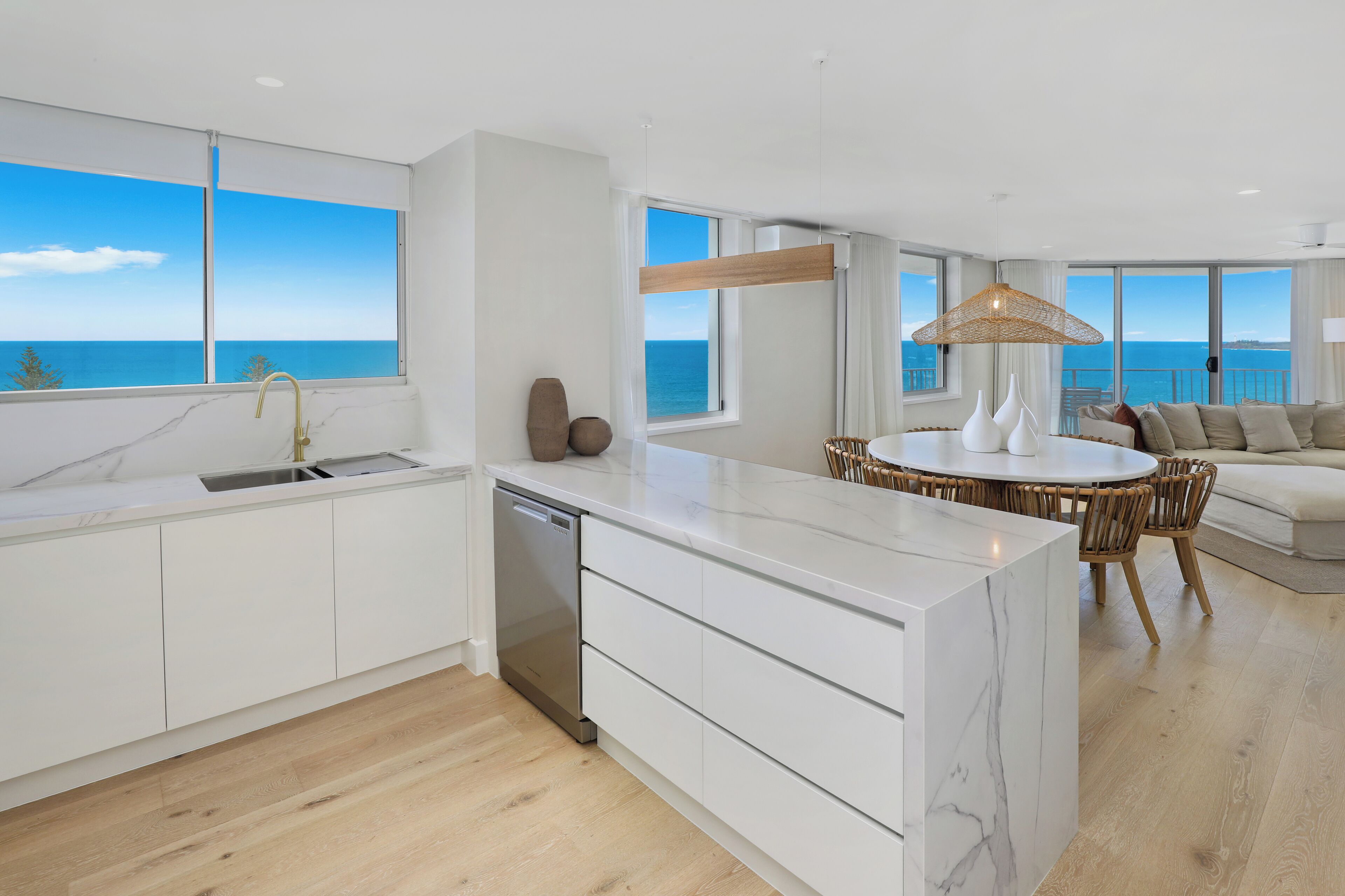 A modern kitchen with a view of the sea, a dining area with a round table, and a living room with a sofa and a large window.