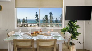 Un coin repas avec une table dressée pour quatre personnes, une plante en pot et une vue sur l’océan et les arbres, visible par une fenêtre.