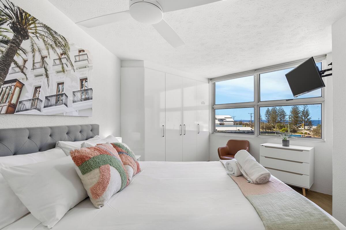 A modern bedroom with a large bed, a ceiling fan, a window with a view, and a flat-screen TV mounted on the wall.