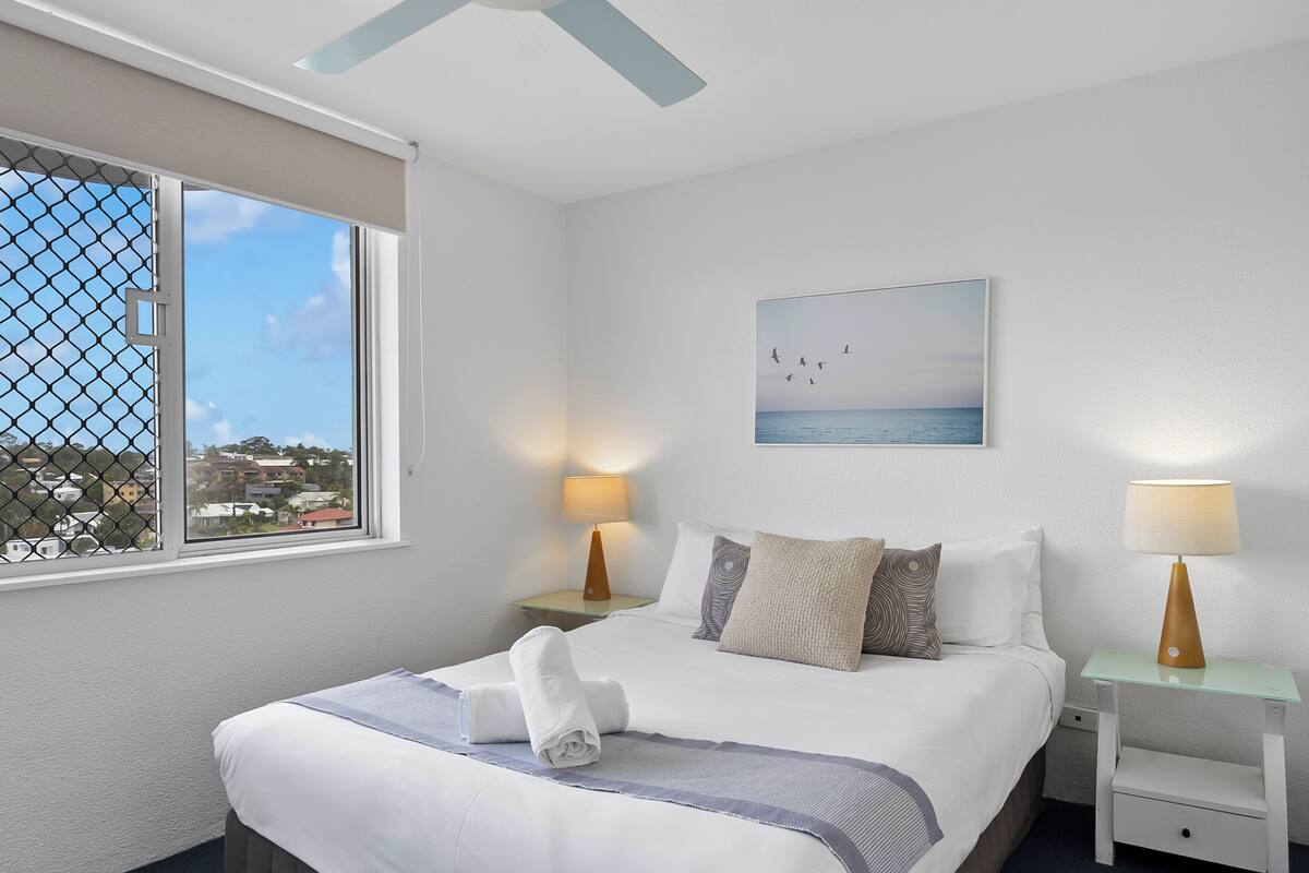 A hotel room with a bed, two bedside tables, a window with a view, and a painting on the wall.