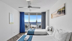 Une chambre d’hôtel avec deux lits, un ventilateur de plafond, une grande fenêtre donnant sur la plage et un tableau au mur.