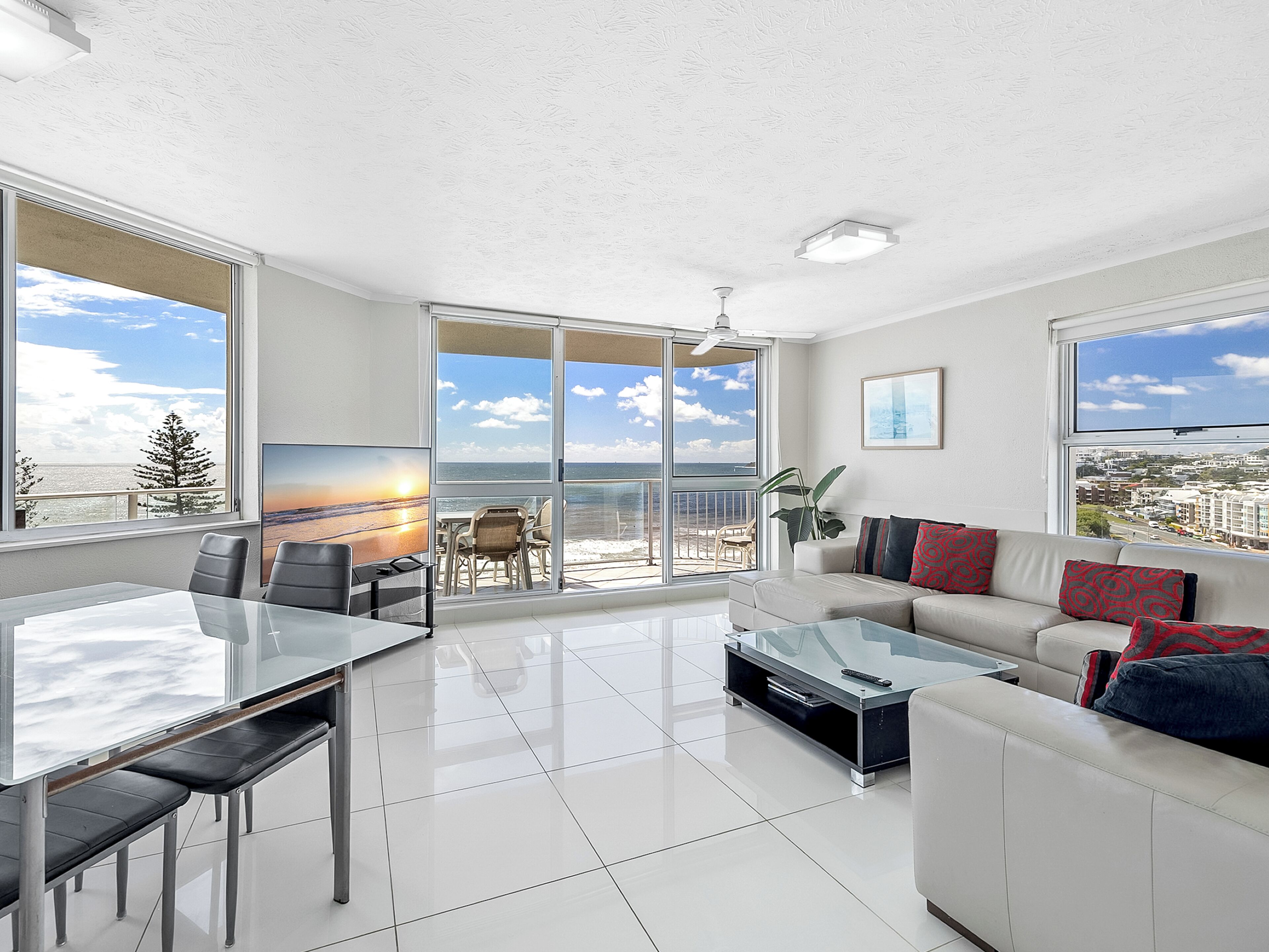 A modern living room with a glass dining table, a sofa with patterned cushions, and a large window offering a view of the ocean.