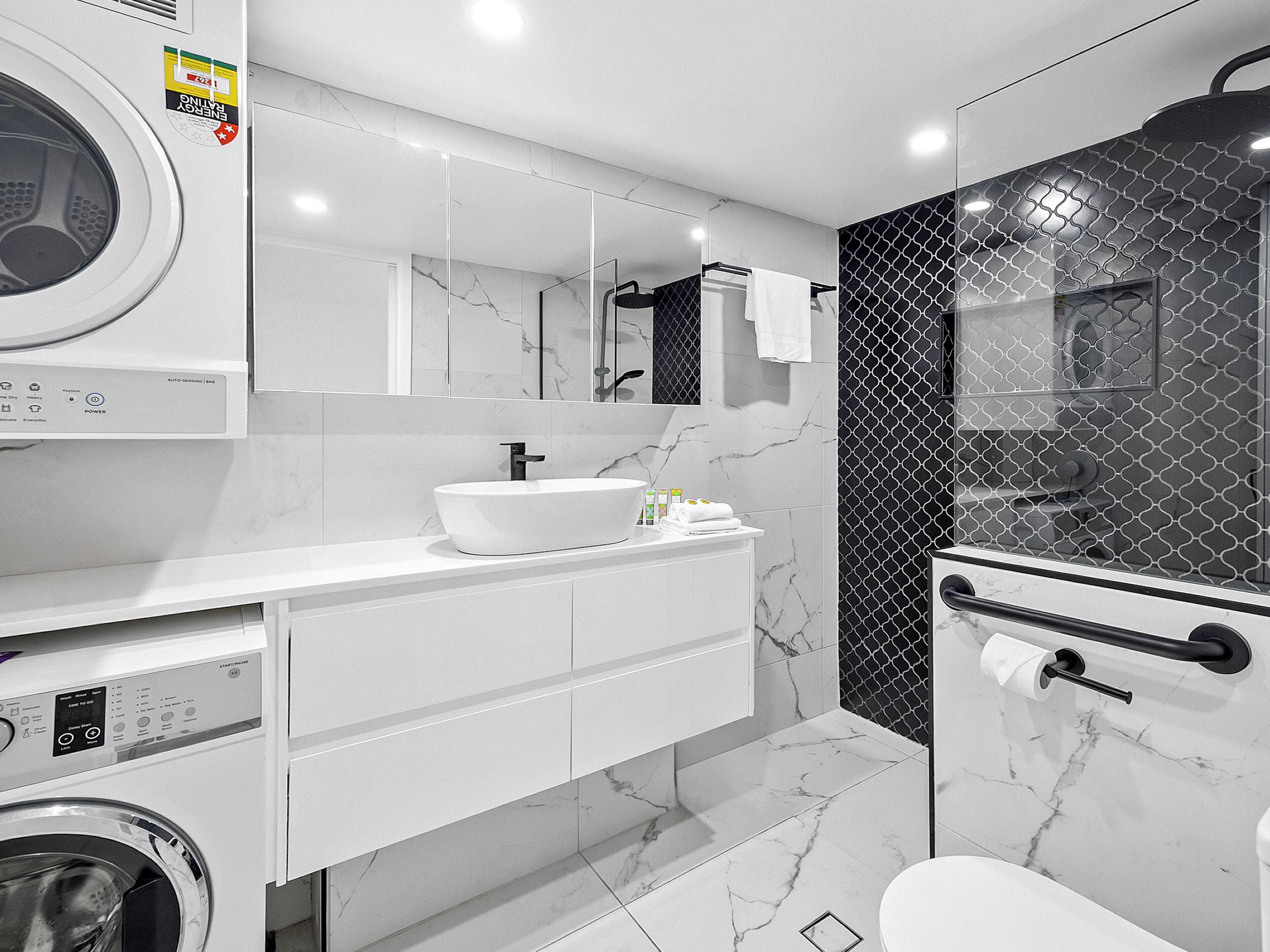 A modern bathroom with a built-in washing machine, a large mirror, a white sink, and a shower area.