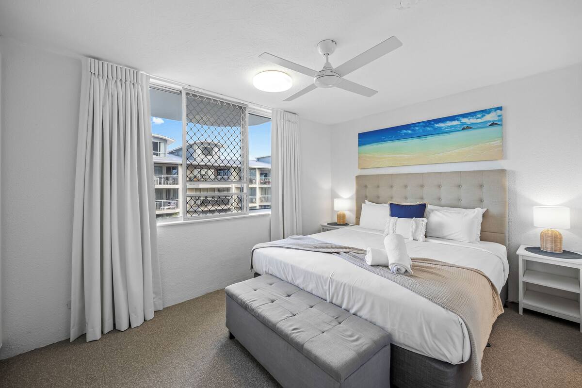 A hotel room with a large bed, a ceiling fan, a window with curtains, a painting of a beach, and a bedside table with a lamp.