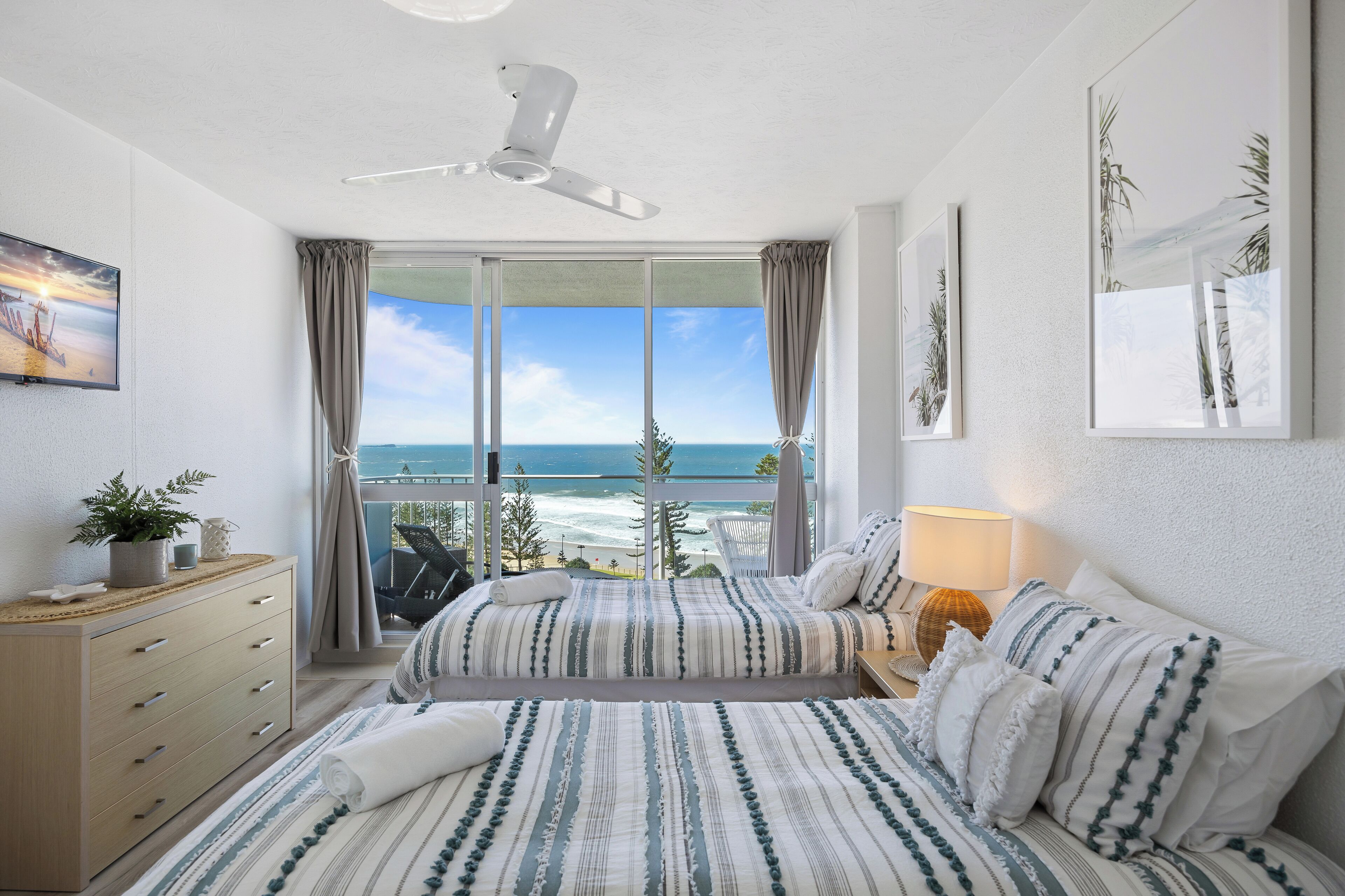 A bedroom with a large bed, a dresser, a nightstand with a lamp, and a view of the ocean through a sliding glass door.