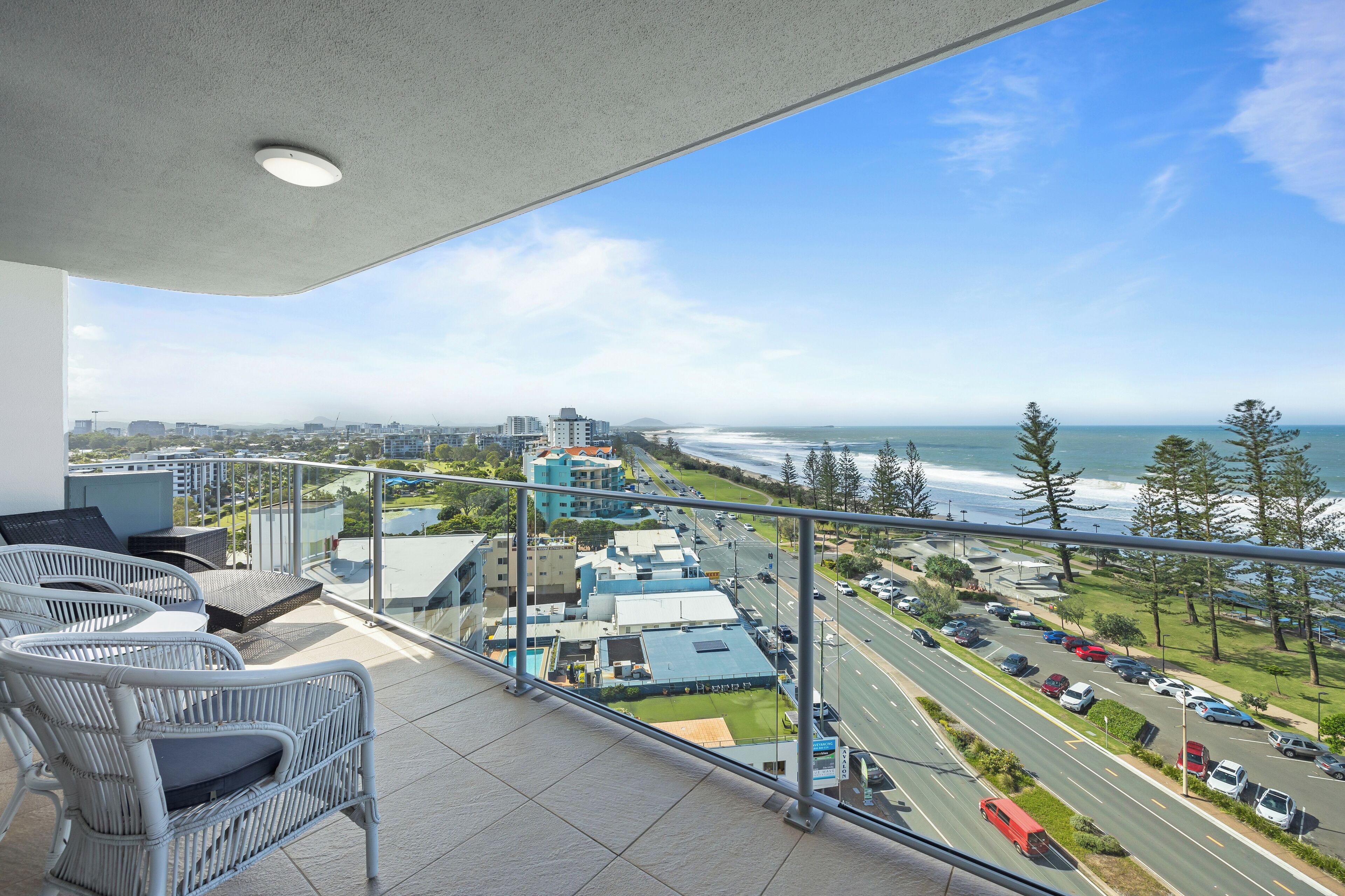 A balcony with wicker furniture overlooking a coastal road and buildings.