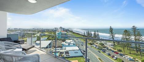 A balcony with wicker furniture overlooking a coastal road and buildings.