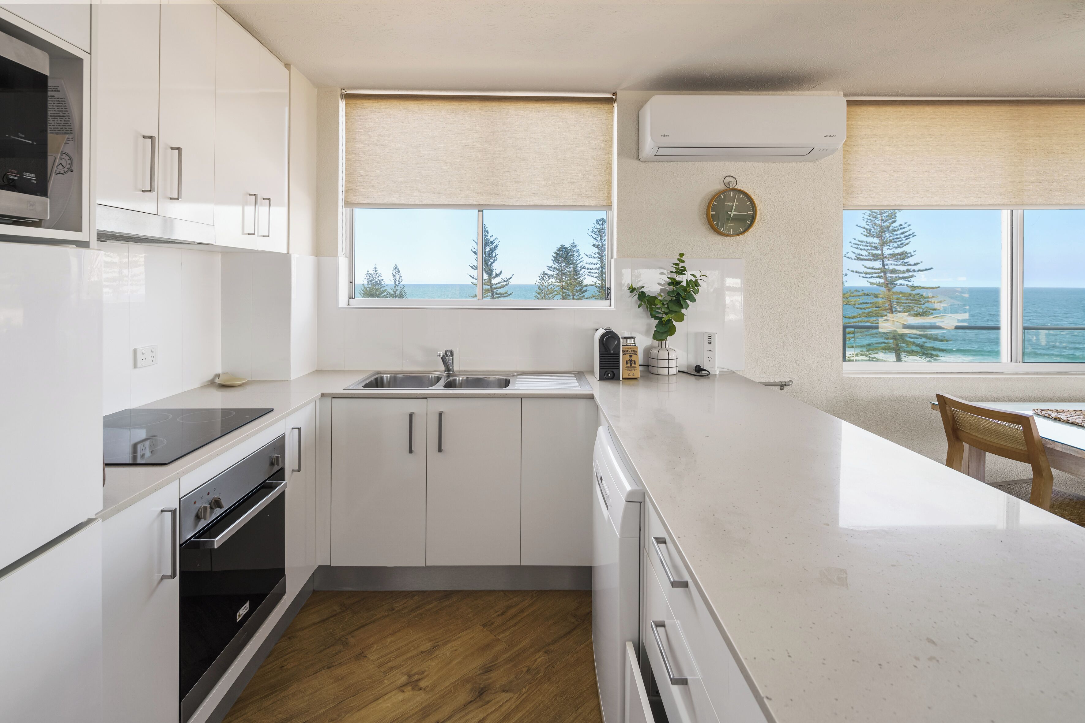A modern kitchen with white cabinetry, a built-in oven, a sink, and a large window offering a view of the ocean and trees.