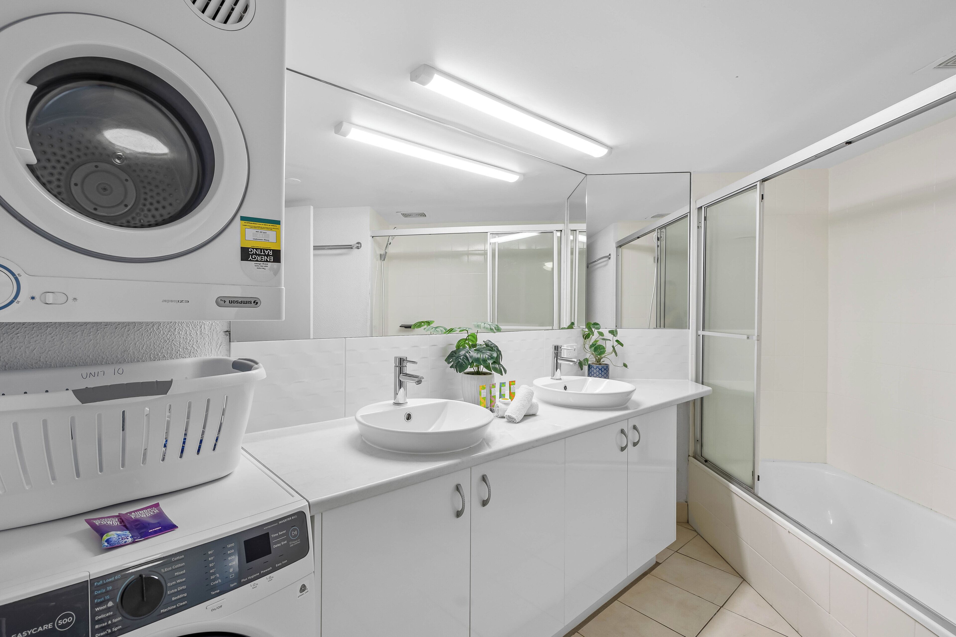 A modern bathroom with a washing machine, two sinks, a large mirror, and a glass shower enclosure.