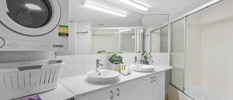 A modern bathroom with a washing machine, two sinks, a large mirror, and a glass shower enclosure.