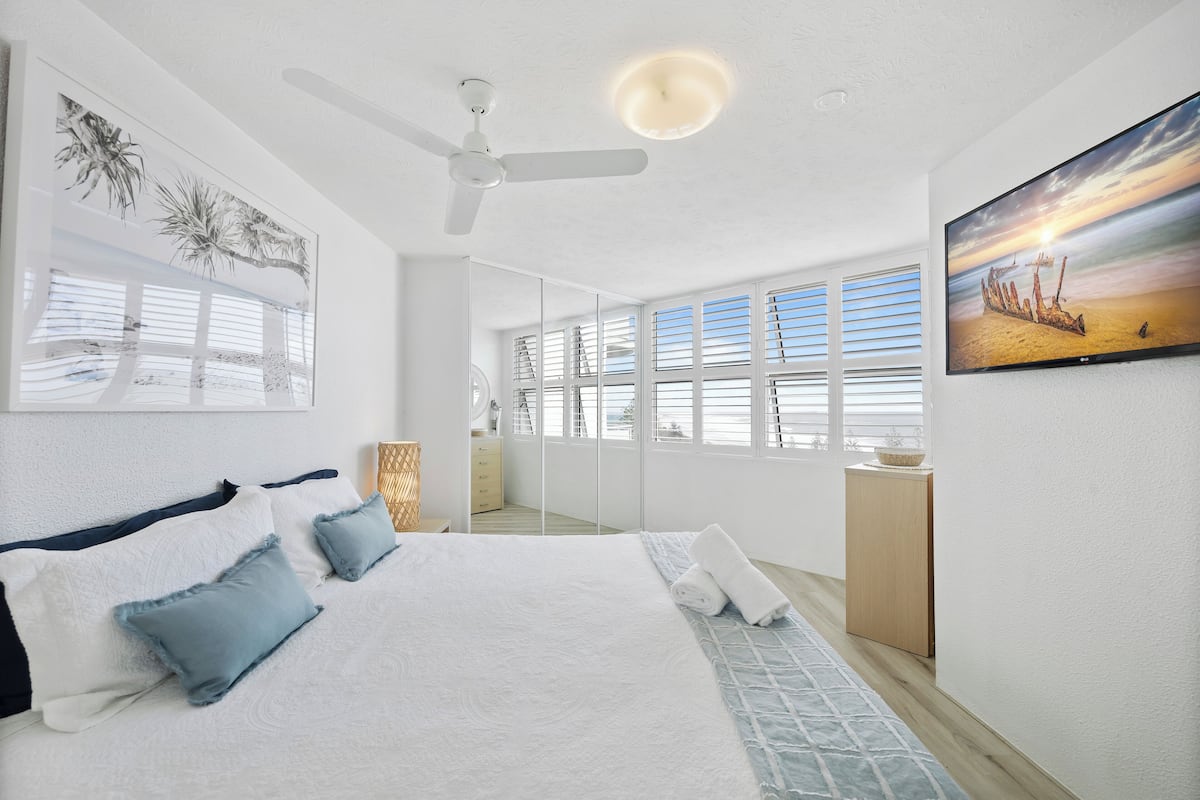 A bedroom with a large bed, a ceiling fan, a painting of a sunset, and a view of the beach through the window.
